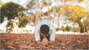 A man kneeling in prayer on a ground covered with autumn leaves, with trees in the background, symbolizing humility, devotion, and seeking blessings through obedience.