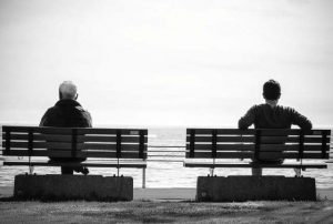 Two people resting on separate benches facing the ocean in a black-and-white scene, with a grassy foreground and a railing in view