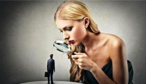 Woman closely examining a tiny man with a magnifying glass, symbolizing critical scrutiny or over-analysis.