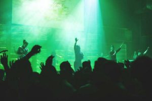 A live worship concert scene with performers holding microphones on stage illuminated by green lighting effects, while an engaged audience raises hands in the foreground.